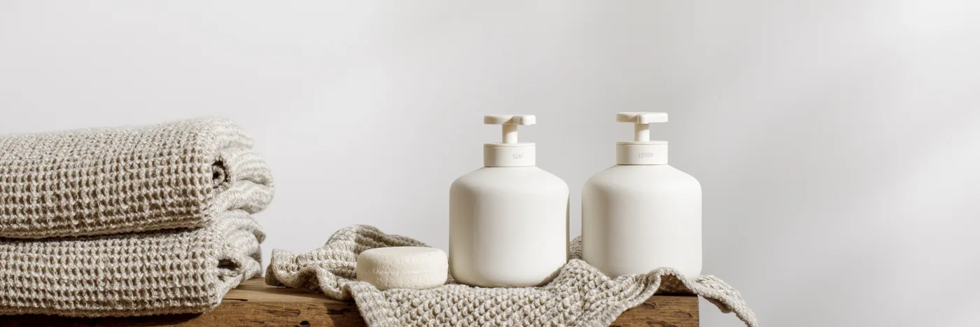 Two unbranded white pump bottles and a folded linen towel on a weathered oak block, the wash and lotion rungs of a fragrance layering routine.