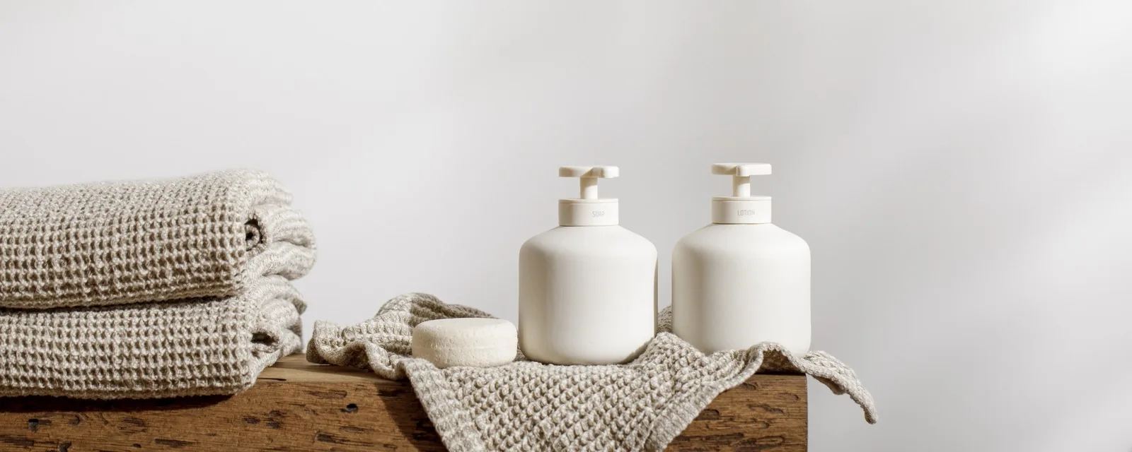 Two unbranded white pump bottles and a folded linen towel on a weathered oak block, the wash and lotion rungs of a fragrance layering routine.
